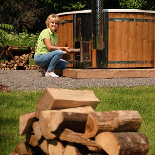 Woman in a green shirt and blue jeans adding firewood to a wood-fired hot tub heater in a garden setting