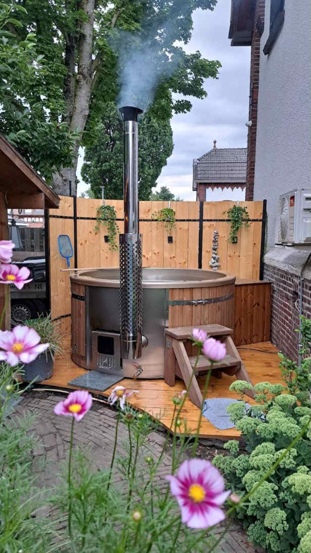 Outdoor view of a wood fired hot tub with metal chimney, wooden steps, and floral garden in foreground