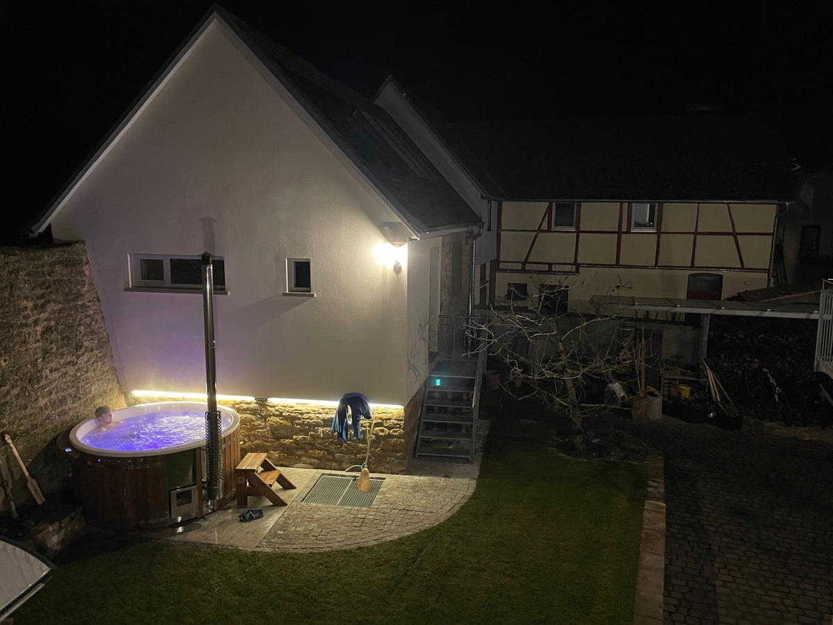 Nighttime view of illuminated outdoor hot tub beside stone and plaster house with adjacent garden and wooden stairs