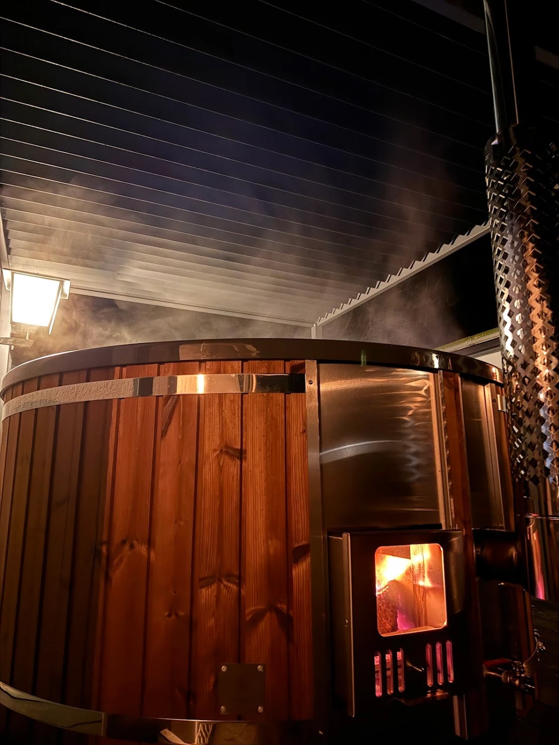 Night view of wood fired hot tub with visible fire through stove window and steam rising under roofed outdoor area