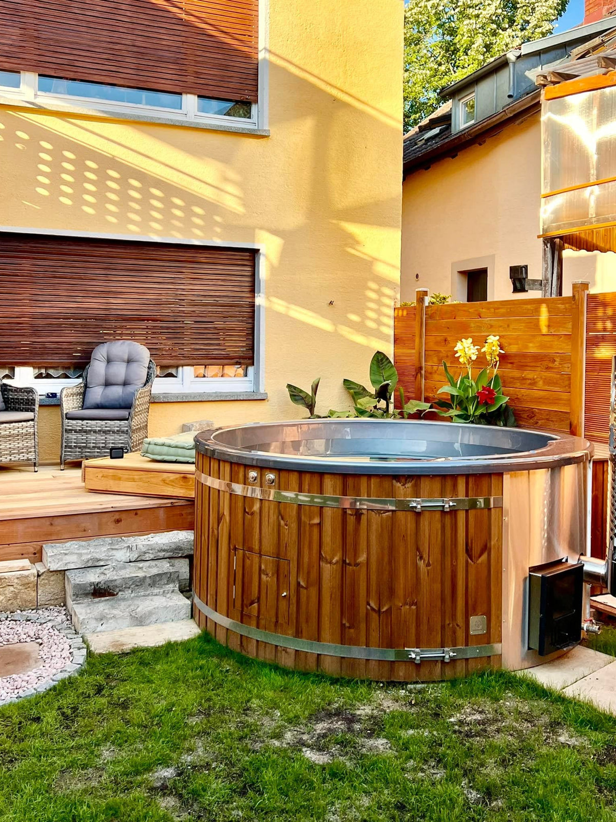 Gardenvity wood fired hot tub in urban garden beside yellow house with patio furniture.