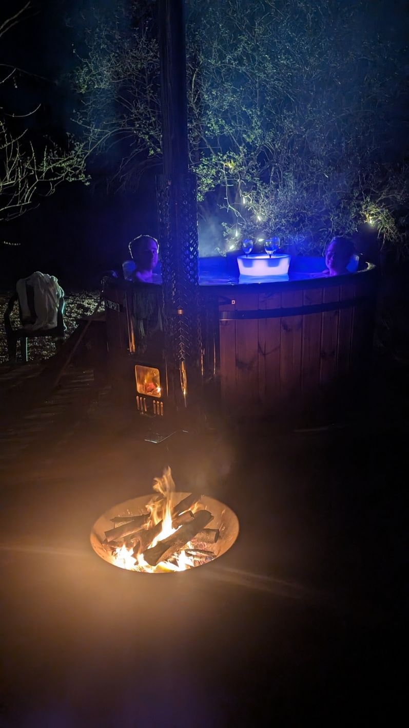 Nighttime view of two people relaxing in a wood fired hot tub near a glowing fire pit and trees in background
