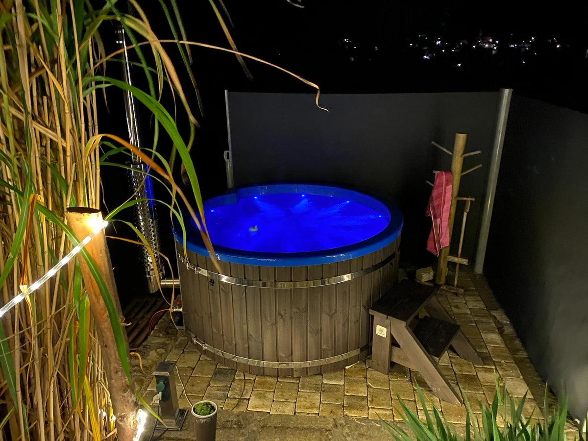 Night view of round wooden hot tub with blue LED lighting on stone tile patio, small wooden steps, and outdoor shower area
