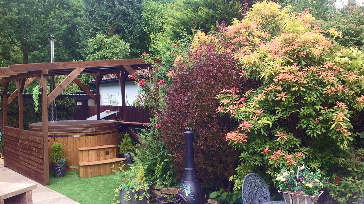 Garden patio with wooden pergola, hot tub, lush shrubs, outdoor chiminea, and metal table with chairs.