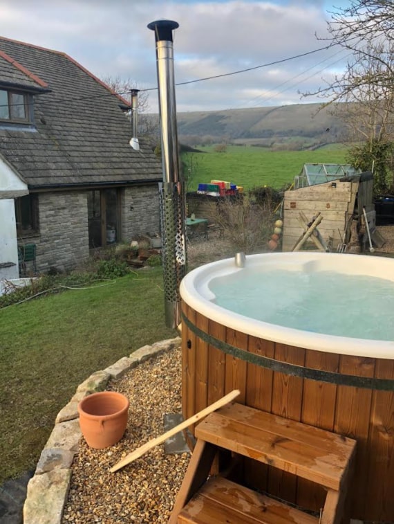 Outdoor view of round wood fired hot tub with integrated stainless steel stove and wooden steps beside stone house