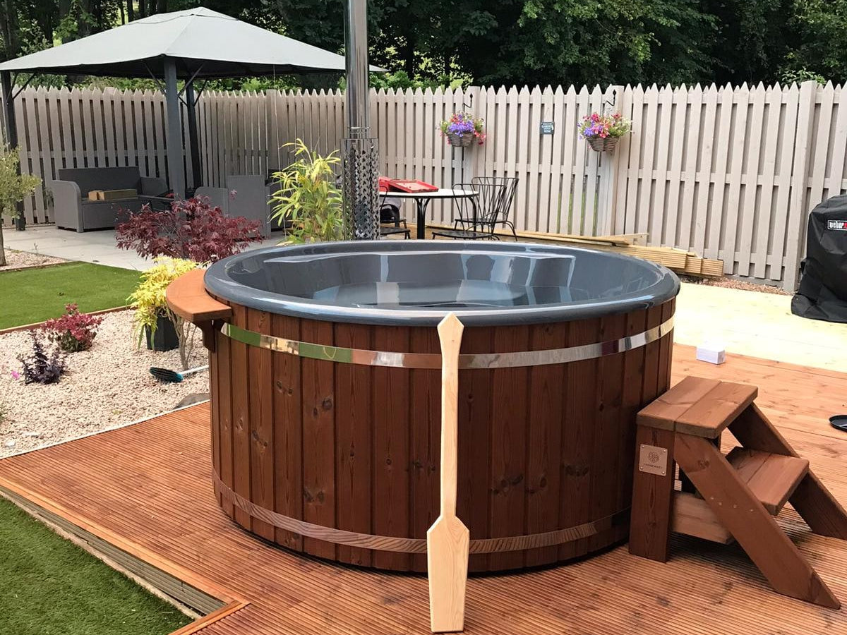 Outdoor view of round wooden hot tub with metal bands, chimney, wooden steps, and paddle on wooden deck area
