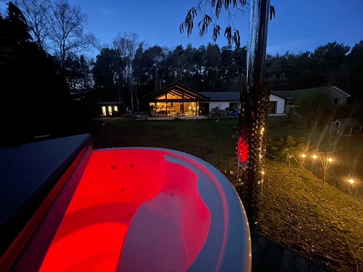 Nighttime outdoor view of illuminated wood fired hot tub with red lighting near a lit modern cabin in forest setting