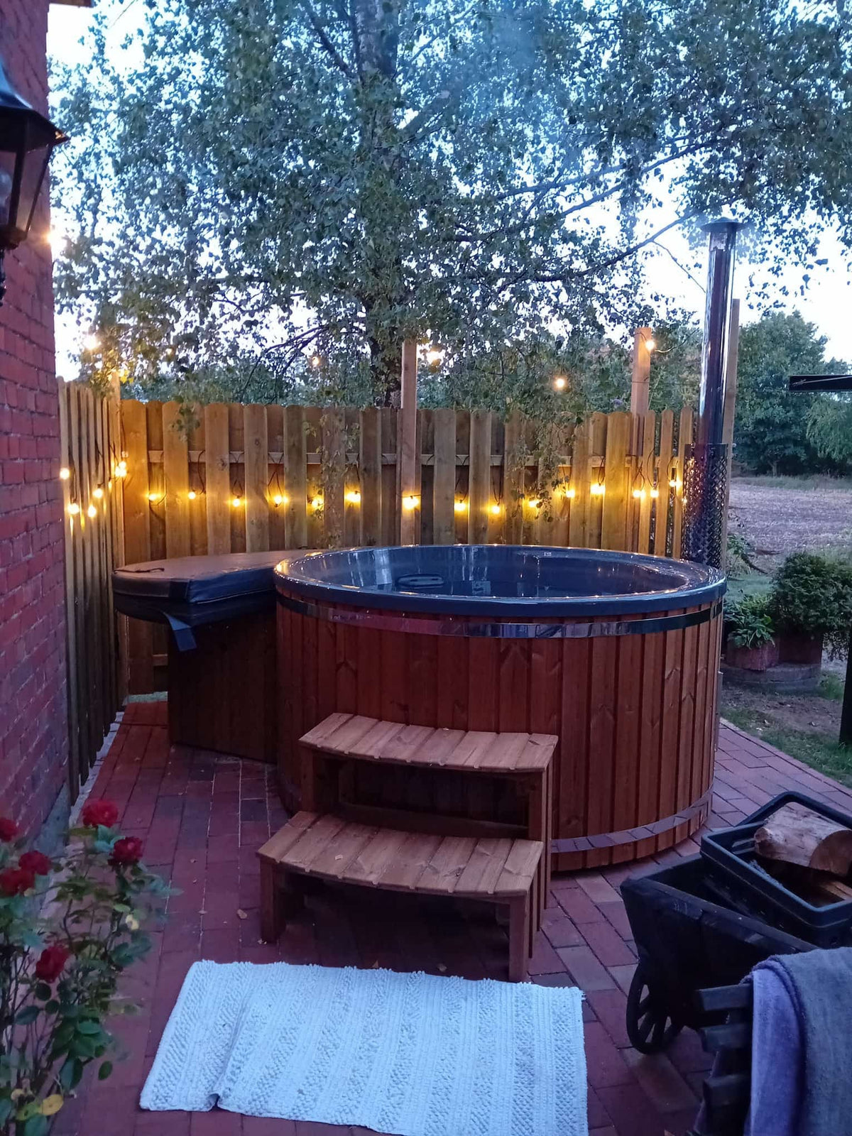 Outdoor view of a wooden hot tub with two-step wooden stairs, string lights on fence, and brick patio at dusk