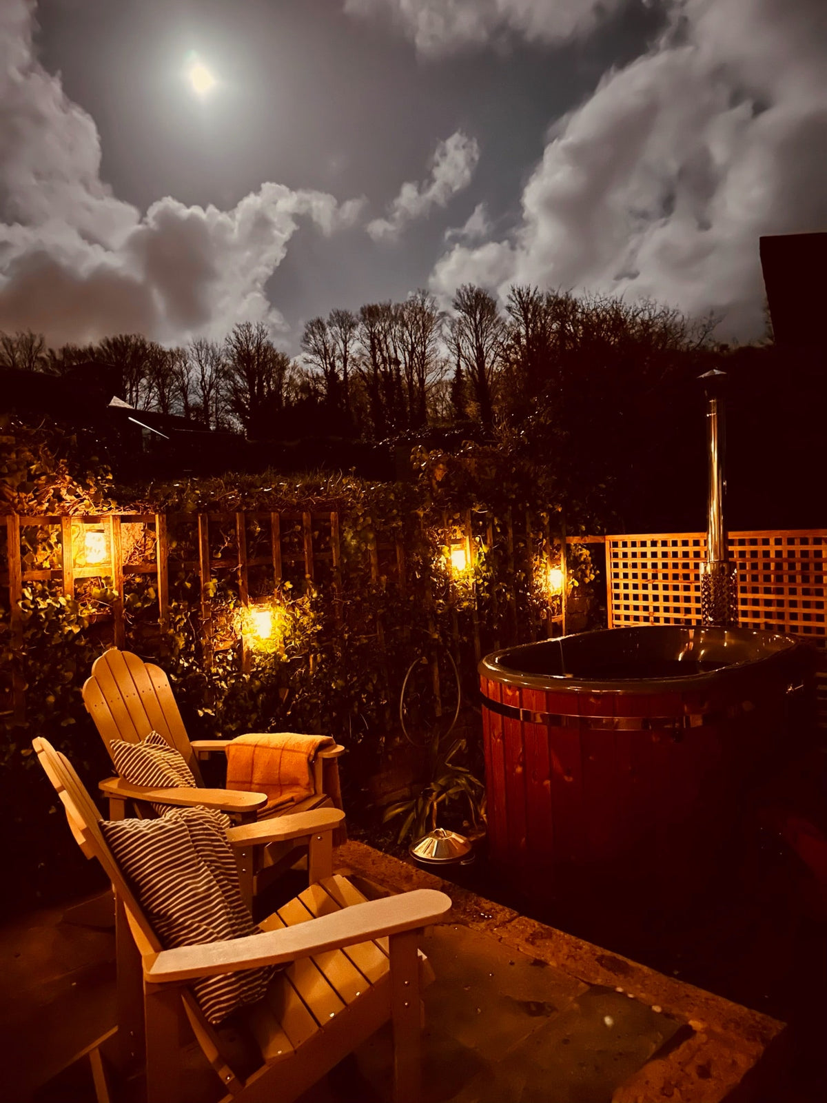 Night view of outdoor wooden hot tub with chimney, Adirondack chairs with cushions, and string lights under a cloudy moonlit sky