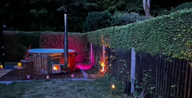 Outdoor wood fired hot tub with lit stove and colorful garden lighting surrounded by greenery and wooden fence at dusk