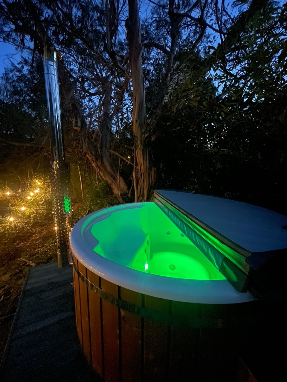 Outdoor view of wood fired hot tub lit with green LED lights and surrounded by trees and decorative string lights at dusk